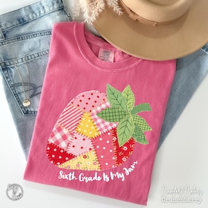 Può includere: T-shirt rosa con un design a patchwork di fragola e la scritta "Sixth Grade Is My Jam". La fragola è realizzata con vari tessuti fantasia rossi, rosa e gialli. Sono attaccate foglie verdi. Un cappello beige e jeans blu sono sullo sfondo.