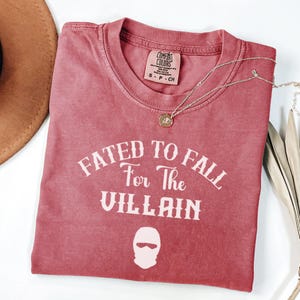 May include: A faded red Comfort Colors t-shirt featuring the text "Fated to Fall For The Villain" in white, with a ski mask graphic. A gold pendant necklace and a brown hat are also visible.