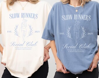 May include: Two women wearing oversized t-shirts with the text "Slow Runners Social Club" in blue lettering. The shirts are in cream and blue colors. The text also includes "Est. 2025".