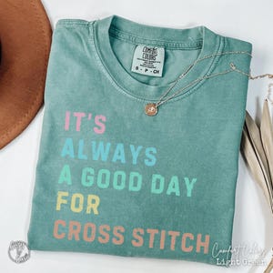May include: A light green t-shirt with the text "It's Always A Good Day For Cross Stitch" in rainbow colours. The shirt is made of a soft, comfortable material and has a distressed look.