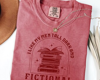 May include: A dusty rose Comfort Colors t-shirt with the text "I LIKE MY MEN TALL DARK AND FICTIONAL" in a circular design with a stack of books. A silver necklace with a pendant is draped on the shirt.