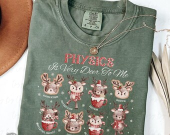 May include: A moss green Comfort Colors t-shirt with the text "PHYSICS Is Very Deer To Me" in red and white. The shirt features cartoon reindeer illustrations with physics terms. A gold necklace is draped across the shirt.