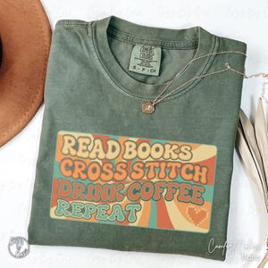 Può includere: Una maglietta Comfort Colors verde muschio con la scritta "READ BOOKS CROSS STITCH DRINK COFFEE REPEAT" in un design retrò. Una collana d'oro è drappeggiata sulla maglietta. La maglietta è di taglia small.