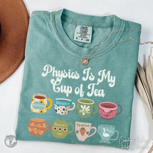 May include: Light green Comfort Colors t-shirt with the phrase "Physics Is My Cup of Tea" and colorful teacup graphics. Each teacup represents a physics concept like "Momentum" and "Inertia". A silver necklace with a pendant is also visible.