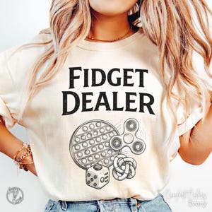 May include: Cream-colored sweatshirt with the text "FIDGET DEALER" in black. Below the text are illustrations of a pop-it toy, a fidget spinner, a dice, and a tangle toy. The shirt is labeled "Comfort Colors Ivory".