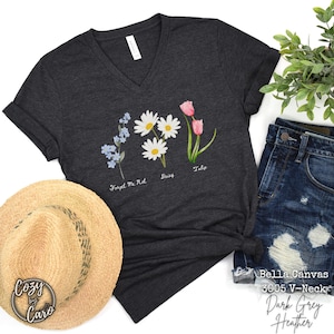 May include: Dark grey heather V-neck t-shirt featuring watercolor illustrations of flowers: forget-me-nots, daisies, and tulips. The shirt has rolled sleeves and is styled with a straw hat and denim shorts. The text "Forget Me Not", "Daisy", and "Tulip" are below the flowers.