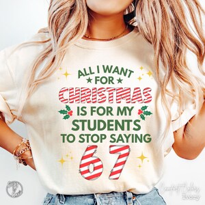 May include: Ivory-colored sweatshirt with the text "ALL I WANT FOR CHRISTMAS IS FOR MY STUDENTS TO STOP SAYING 67". The text is in red and green with star and holly accents. The sweatshirt is a Comfort Colors brand.