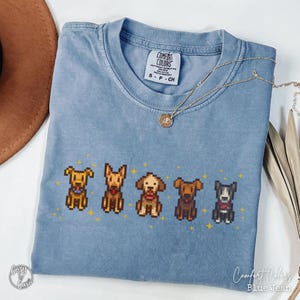 May include: A blue jean Comfort Colors t-shirt featuring a pixelated design of five different dog breeds. The shirt has a crew neck and short sleeves. The text "Comfort Colors" and "Blue Jean" are visible. A gold necklace is draped across the shirt.
