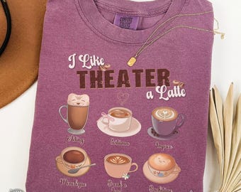 May include: A berry-colored t-shirt with the phrase "I Like Theater a Latte" and coffee cup illustrations. The shirt has a crew neck and a Comfort Colors tag. A gold necklace and a brown hat are also visible.