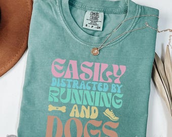 May include: A light green t-shirt with the text "EASILY DISTRACTED BY RUNNING AND DOGS" in a retro font. The text is in pink, blue, green, and orange. The shirt is made by Comfort Colors.