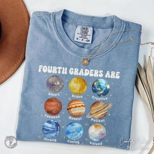 May include: Blue Comfort Colors t-shirt with the text "Fourth Graders Are" and planet graphics. The planets are labeled with positive attributes like "Smart," "Brave," and "Creative." A gold necklace rests on the shirt. The shirt is labeled "Blue Jean."
