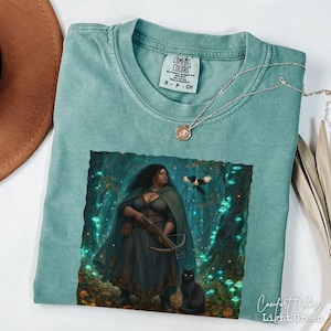 Può includere: Una t-shirt a maniche corte verde chiaro con una stampa grafica di una persona con un mantello scuro che tiene una balestra in una foresta con funghi blu luminosi, un uccello in volo e un gatto nero. Etichetta del marchio Comfort Colors.
