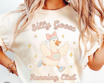 May include: Ivory-colored sweatshirt with the text "Silly Goose Running Club." The design features a cartoon goose wearing a pink running bib with the number 002, a blue headband, and running shoes. The design includes daisies and stars.