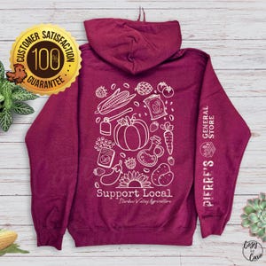 Peut inclure: Un sweat à capuche bordeaux avec un motif blanc représentant un tournesol, des légumes et le texte "Support Local Stardew Valley Agriculture" et "Pierre's General Store".