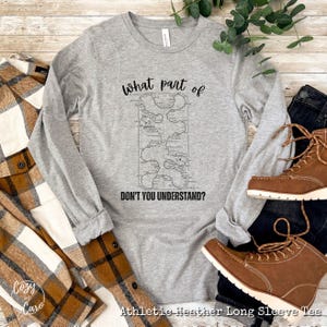 May include: Gray long-sleeve t-shirt with a graphic of a maze and the text "What part of DON'T YOU UNDERSTAND?" printed on the front. The shirt is laid flat on a wooden surface with a plaid shirt and brown boots.