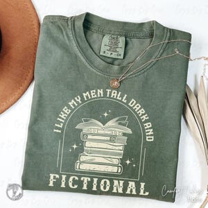 May include: An olive green Comfort Colors t-shirt with the text "I LIKE MY MEN TALL DARK AND FICTIONAL" in white. The graphic features a stack of books. A gold necklace with a pendant rests on the shirt.