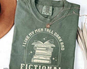 May include: An olive green Comfort Colors t-shirt with the text "I LIKE MY MEN TALL DARK AND FICTIONAL" in white. The graphic features a stack of books. A gold necklace with a pendant rests on the shirt.
