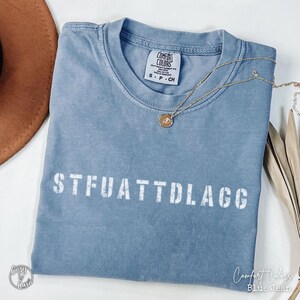 May include: A blue Comfort Colors t-shirt with the text "STFUATTDLAGG" printed in white. A gold necklace with a pendant rests on the shirt. The shirt is folded, and a brown hat is visible in the background.