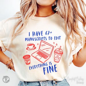 Puede incluir: Sudadera de color marfil con el texto "I HAVE 67+ MANUSCRIPTS TO EDIT EVERYTHING IS FINE" en azul y rojo. El diseño incluye un portátil, una taza de café y café helado. La sudadera es de Comfort Colors.