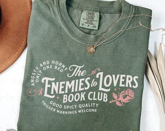 May include: A moss green Comfort Colors t-shirt with the text "The Enemies to Lovers Book Club" and a rose graphic. The shirt also features the phrases "Angsty and Horny Only One Bed", "Good Spicy Quality", and "Trigger Warnings Welcome". A gold necklace is draped on the shirt.