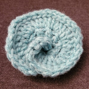 May include: A light blue crocheted flower with a center hole. The flower is made with a single crochet stitch and has a textured appearance.