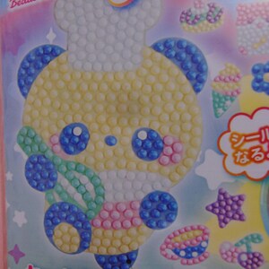 Pu&ograve; includere: Un set di adesivi colorati con un orso chef dei cartoni animati fatto di perline gialle, blu, rosa e bianche. Il set include vari disegni artistici con perline come coni gelato, stelle e ciliegie. Il testo "Beads Ant Sticken" &egrave; visibile.