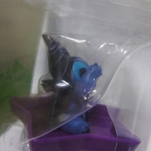 May include: A small, blue and purple figurine of a cartoon character, possibly a dragon or similar creature, is encased in clear plastic. The figurine has large blue eyes and is seated on a purple platform.