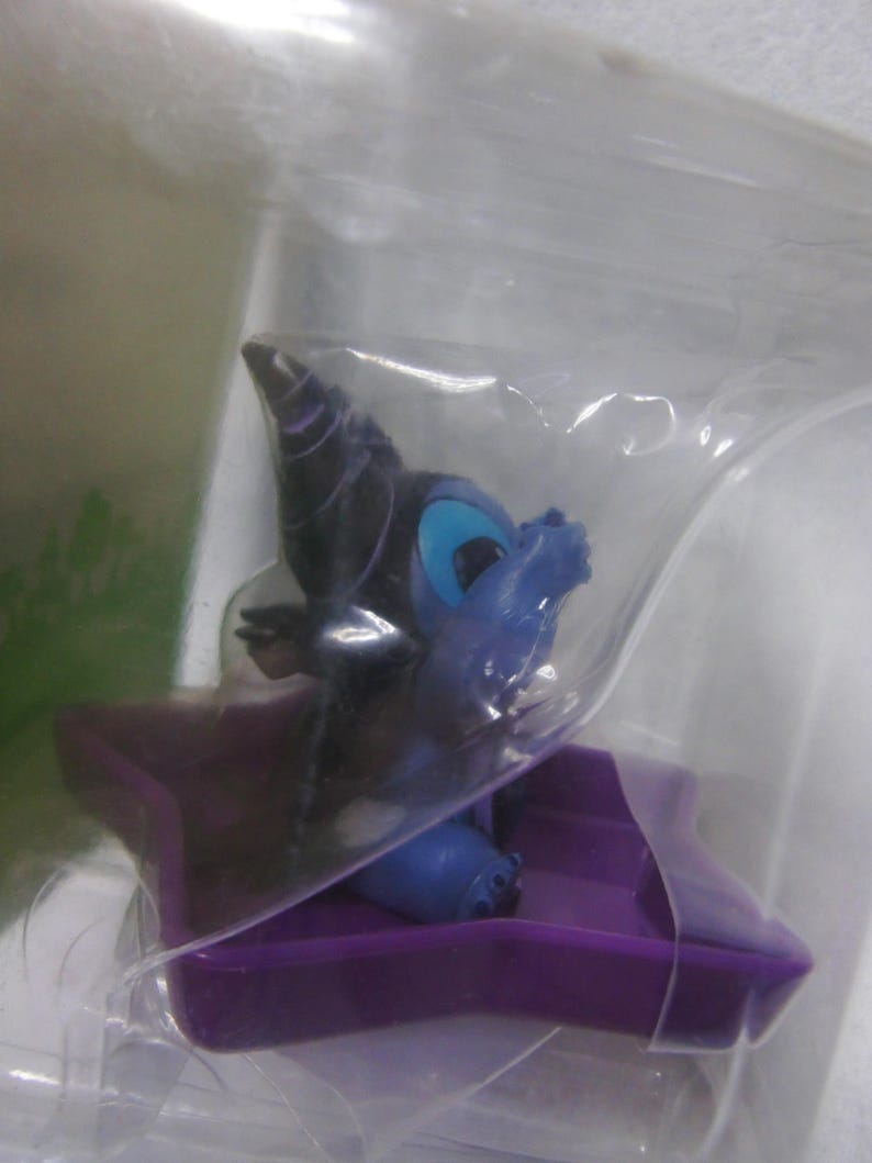 May include: A small, blue and purple figurine of a cartoon character, possibly a dragon or similar creature, is encased in clear plastic. The figurine has large blue eyes and is seated on a purple platform.