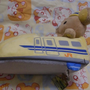 Pu&ograve; includere: Un giocattolo di peluche a forma di treno proiettile giallo con un orso marrone che indossa un cappello da conduttore seduto sopra. Il treno ha una striscia blu e il testo "JR" sul lato. Il giocattolo ha un'etichetta che dice "Tokyo" e "Shinkansen".