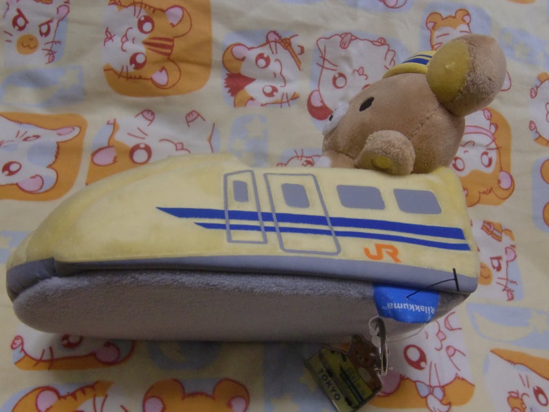 Pu&ograve; includere: Un giocattolo di peluche a forma di treno proiettile giallo con un orso marrone che indossa un cappello da conduttore seduto sopra. Il treno ha una striscia blu e il testo "JR" sul lato. Il giocattolo ha un'etichetta che dice "Tokyo" e "Shinkansen".