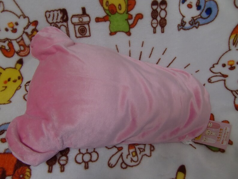 Puede incluir: Una almohada de felpa rosa con forma de cabeza de oso. La almohada tiene una textura suave y aterciopelada y est&aacute; sobre un fondo blanco con personajes de dibujos animados impresos en ella. La almohada tiene una etiqueta adjunta que dice "まめねこ" en japon&eacute;s.