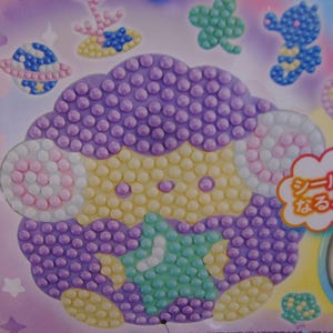 Pu&ograve; includere: Un foglio di adesivi con una pecora dei cartoni animati fatta di perline viola, gialle e bianche. Altri adesivi includono un pianeta, un cavalluccio marino e un trifoglio. Il testo "ads Ant Sticken" &egrave; visibile in alto.