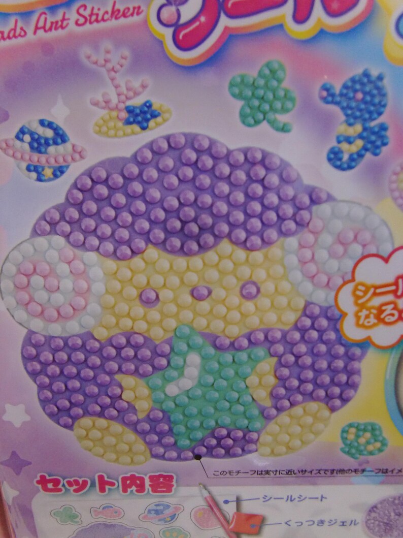 Pu&ograve; includere: Un foglio di adesivi con una pecora dei cartoni animati fatta di perline viola, gialle e bianche. Altri adesivi includono un pianeta, un cavalluccio marino e un trifoglio. Il testo "ads Ant Sticken" &egrave; visibile in alto.