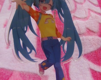 May include: A clear acrylic charm featuring a colorful anime character with long blue pigtails, wearing a yellow shirt, blue pants, and pink shoes. The charm has a silver chain and a base with the name "Miku".