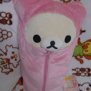 Puede incluir: Un saco de dormir de felpa rosa con una cremallera. El saco de dormir tiene forma de oso y tiene una cara blanca con ojos negros y una nariz negra. El saco de dormir est&aacute; sobre una manta blanca con otros peluches.