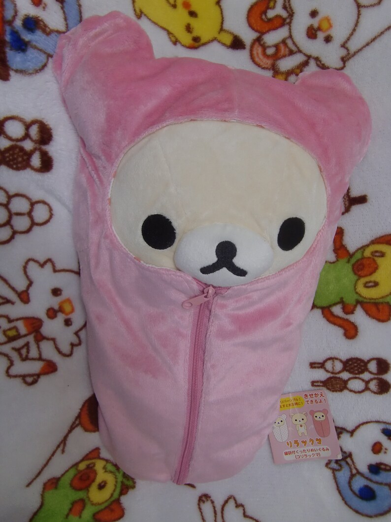 Puede incluir: Un saco de dormir de felpa rosa con una cremallera. El saco de dormir tiene forma de oso y tiene una cara blanca con ojos negros y una nariz negra. El saco de dormir est&aacute; sobre una manta blanca con otros peluches.