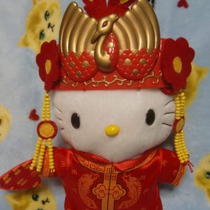 May include: A plush Hello Kitty doll wearing a red and gold traditional Chinese wedding outfit. The outfit includes a decorative headdress with a golden bird, a red jacket with dragon embroidery, and dangling yellow beaded accents.