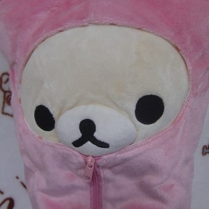 Puede incluir: Una almohada de felpa rosa con una cremallera en la parte delantera. La almohada tiene forma de oso con una chaqueta con capucha rosa. El oso tiene una cara blanca con ojos negros y una nariz negra.