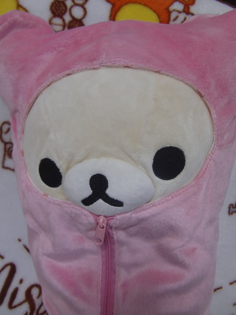 Puede incluir: Una almohada de felpa rosa con una cremallera en la parte delantera. La almohada tiene forma de oso con una chaqueta con capucha rosa. El oso tiene una cara blanca con ojos negros y una nariz negra.