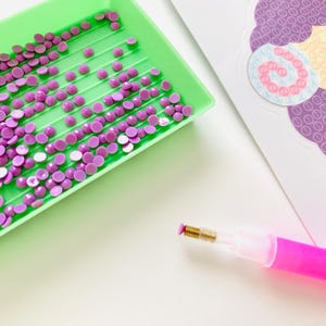 Pu&ograve; includere: Primo piano di materiali per pittura con diamanti. Un vassoio verde contiene numerosi strass rotondi viola. Una penna rosa con punta dorata &egrave; posizionata vicino al vassoio. Una carta decorativa con un disegno viola &egrave; sullo sfondo.