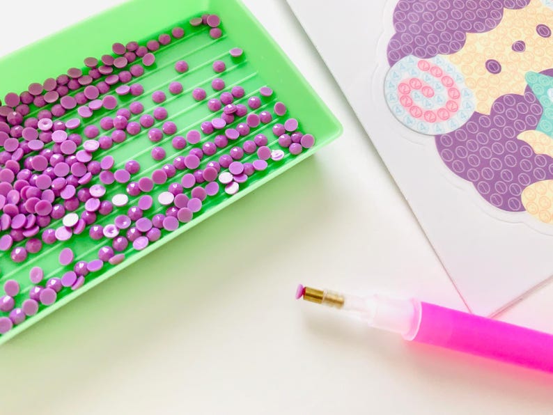 Pu&ograve; includere: Primo piano di materiali per pittura con diamanti. Un vassoio verde contiene numerosi strass rotondi viola. Una penna rosa con punta dorata &egrave; posizionata vicino al vassoio. Una carta decorativa con un disegno viola &egrave; sullo sfondo.