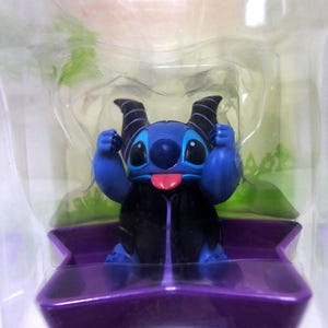 May include: A blue figurine of a cartoon character with large eyes, a pink tongue, and black horns. The figurine is in a clear plastic package, sitting on a purple base. The character is wearing a black cape.