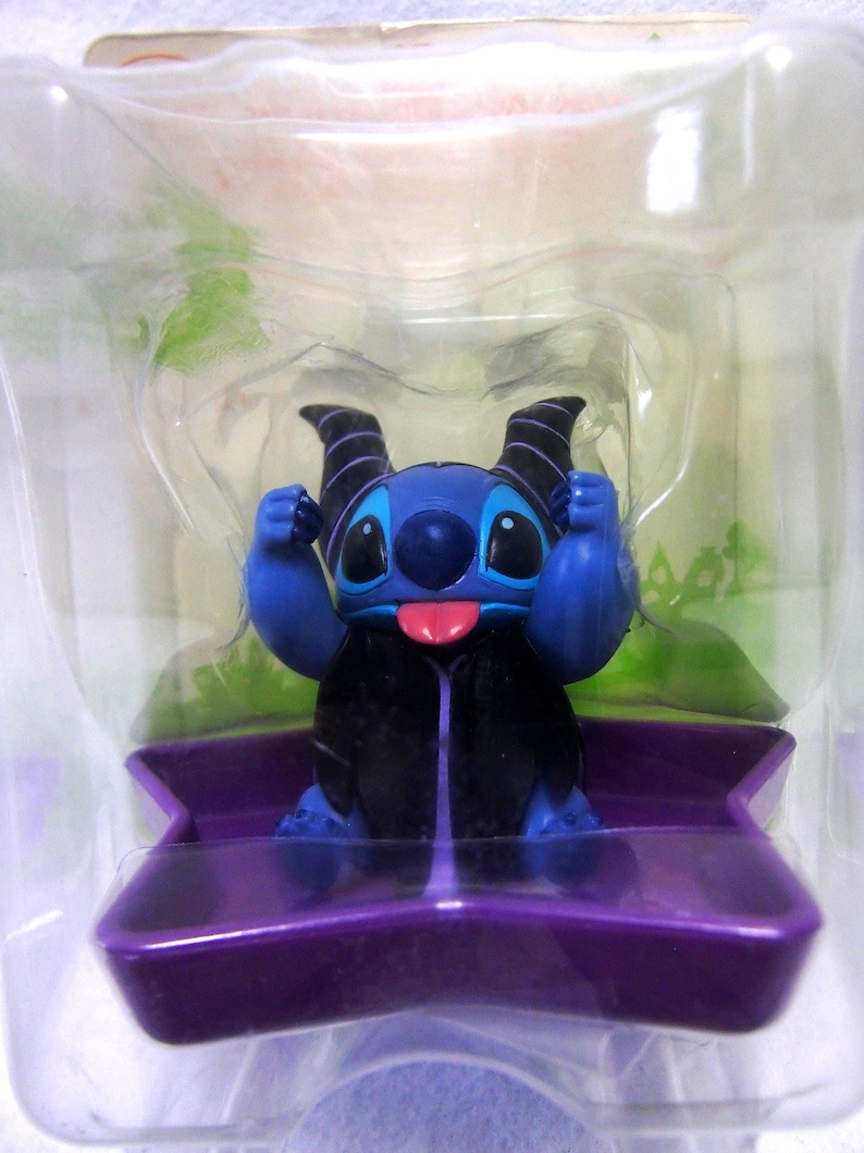 May include: A blue figurine of a cartoon character with large eyes, a pink tongue, and black horns. The figurine is in a clear plastic package, sitting on a purple base. The character is wearing a black cape.