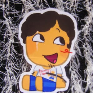 Puede incluir: Una pegatina con un personaje de dibujos animados con pelo casta&ntilde;o, cara naranja y pantalones cortos azules. El personaje sonr&iacute;e con la boca roja y una l&iacute;nea roja en la nariz. La camiseta dice "BLAKE 301".