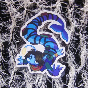 Puede incluir: Una pegatina con un personaje de dibujos animados con cuerpo azul, pelo oscuro y una luz amarilla brillante. El personaje tiene una cola de pez y lleva una bufanda verde y roja. La pegatina est&aacute; sobre un fondo blanco y negro texturizado.