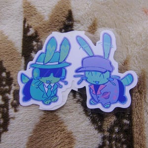Puede incluir: Dos pegatinas con conejos de dibujos animados con trajes elegantes. Un conejo lleva un traje turquesa, gafas de sol y un sombrero. El otro conejo lleva una chaqueta y gorra moradas. Ambos tienen cuerpos turquesas y est&aacute;n sonriendo.