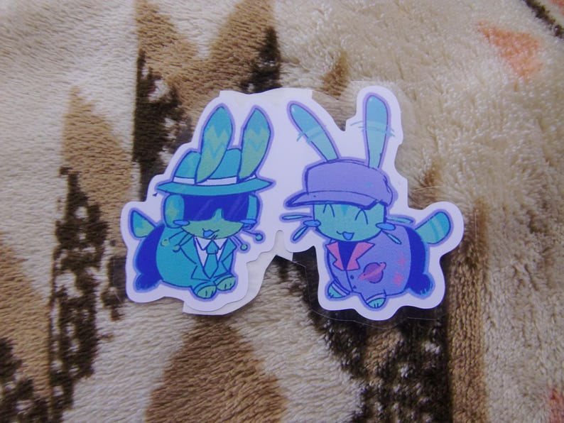 Puede incluir: Dos pegatinas con conejos de dibujos animados con trajes elegantes. Un conejo lleva un traje turquesa, gafas de sol y un sombrero. El otro conejo lleva una chaqueta y gorra moradas. Ambos tienen cuerpos turquesas y est&aacute;n sonriendo.