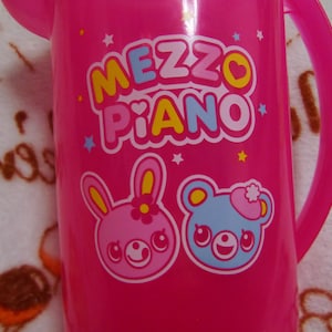 Op de afbeelding: Een felroze plastic kan met een handvat. De kan toont de tekst "MEZZO PIANO" in kleurrijke letters, samen met cartoon illustraties van een roze konijn en een blauwe beer. Kleine sterren zijn verspreid over het ontwerp.