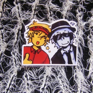 Puede incluir: Una pegatina con dos personajes de dibujos animados. Uno tiene el pelo amarillo, una diadema roja y una camisa roja, haciendo una se&ntilde;al de pulgar hacia arriba. El otro lleva un traje y sombrero negros con gafas de sol. Ambos sonr&iacute;en.