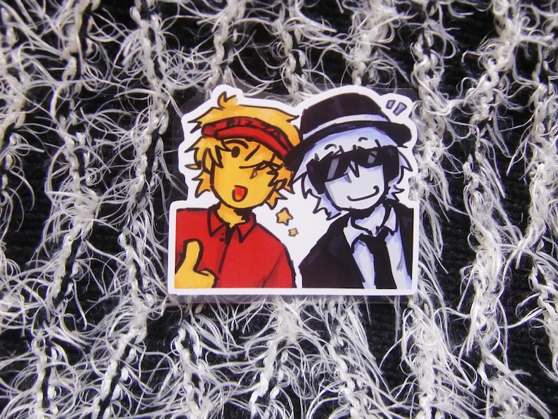 Puede incluir: Una pegatina con dos personajes de dibujos animados. Uno tiene el pelo amarillo, una diadema roja y una camisa roja, haciendo una se&ntilde;al de pulgar hacia arriba. El otro lleva un traje y sombrero negros con gafas de sol. Ambos sonr&iacute;en.